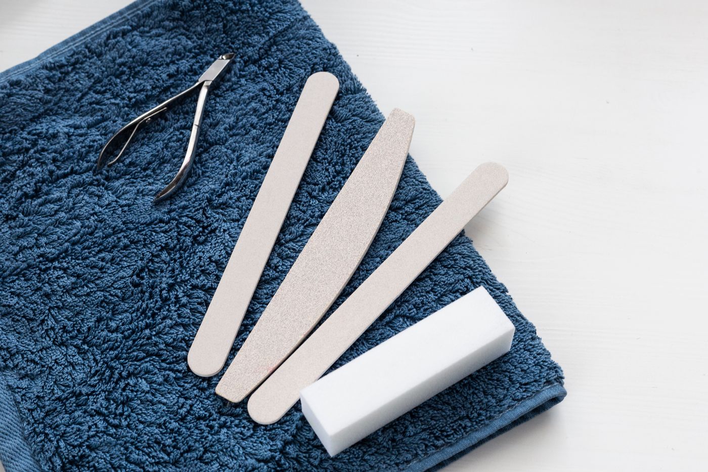 A folded blue terry towel on a white surface carrying cuticle nippers, three beige nail files and a white buffing block.