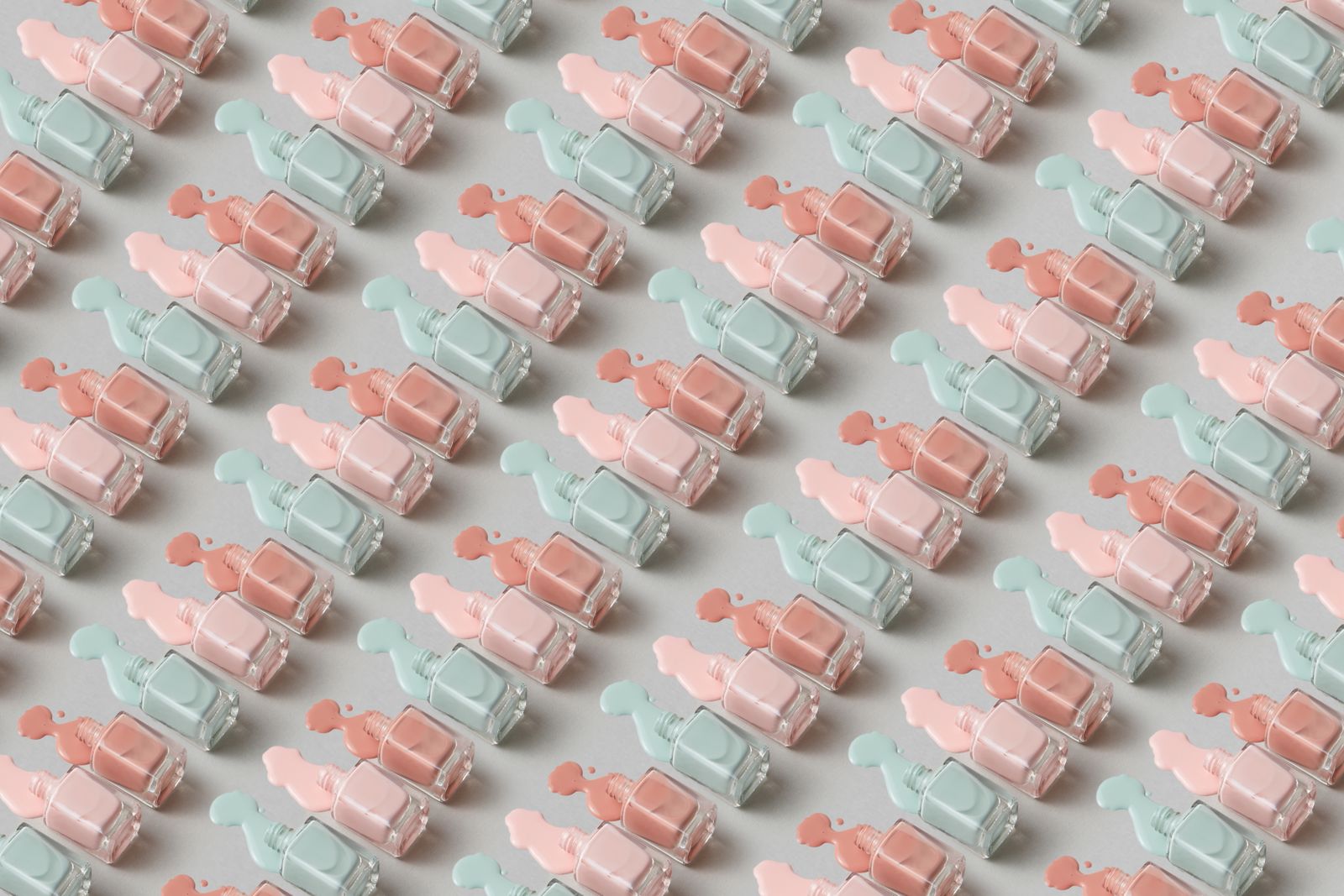 A repeating diagonal pattern of square nail-polish bottles lying on a grey surface in pastel mint, blush pink and peach, each with a small spill of its colour.