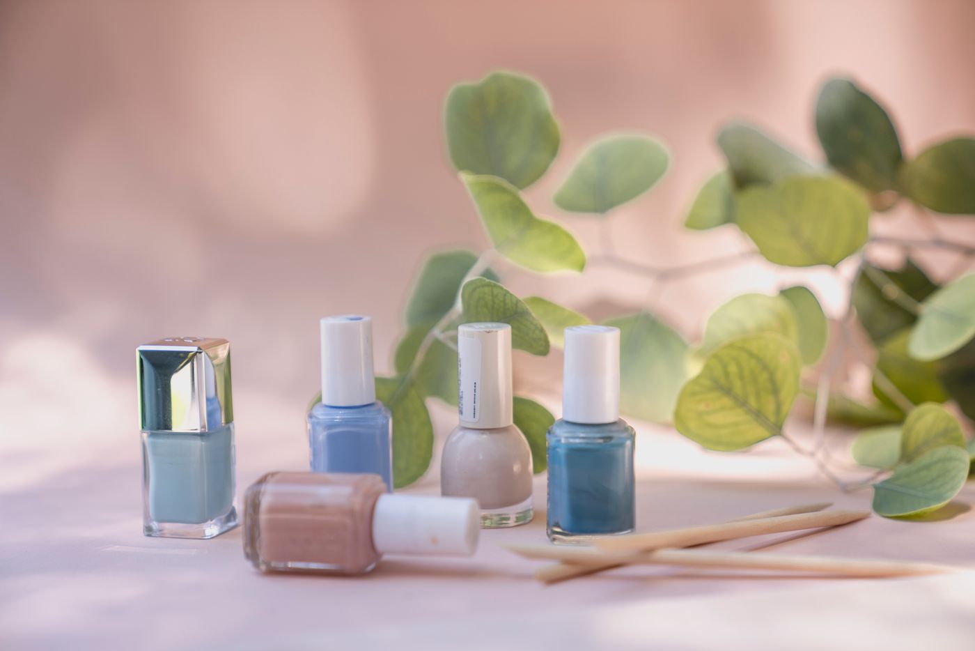 Five pastel nail-polish bottles with orange sticks in front of eucalyptus leaves on a soft peach backdrop.