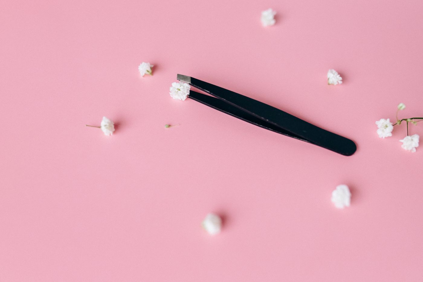Black slant-tip tweezers lying among small white baby's-breath blossoms on a flat pink background.
